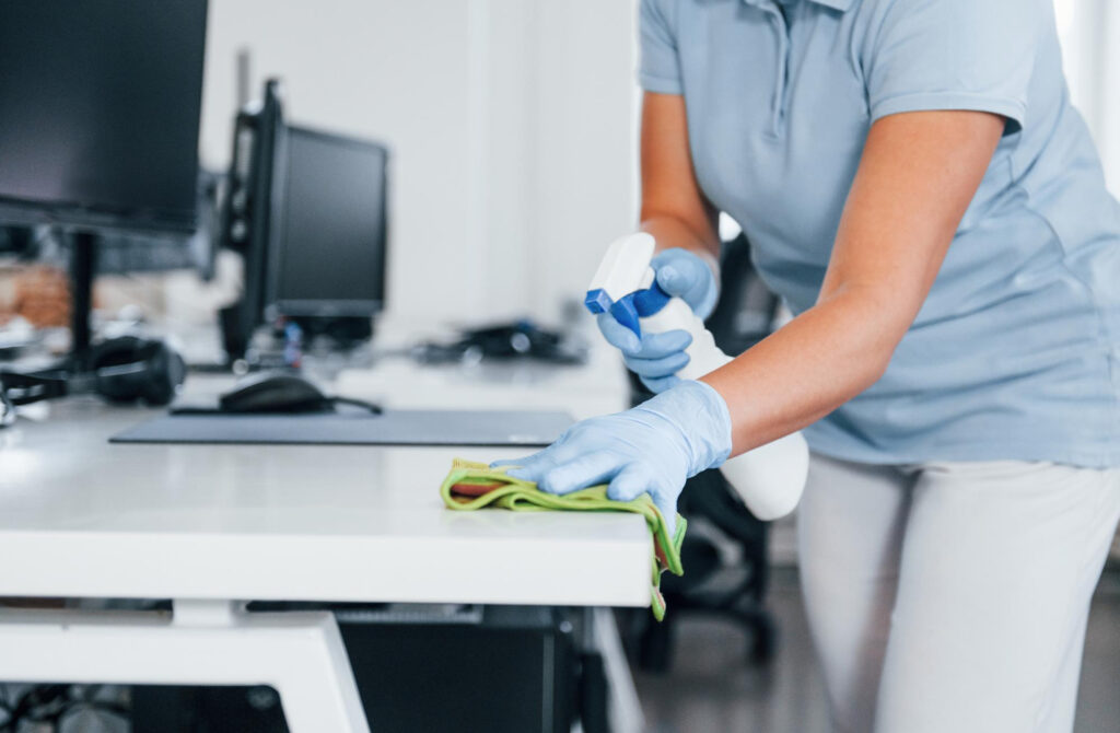 A person wearing gloves cleans an office desk with a cloth and spray bottle, showcasing business cleaning expertise while computers sit in the background—perfect for promoting professional Office Cleaning Services.